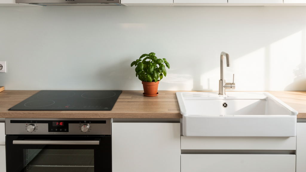 A wide-angle, high-resolution photo of a modern, bright kitchen featuring a wooden countertop between a sleek induction stove and a deep porcelain sink. A small potted basil plant sits in the center. Soft, natural morning light, 8k, photorealistic style.