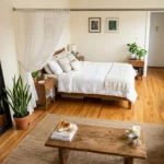 A bright, lived-in studio apartment viewed from above at an angle, showing a bed in command position with a leaning mirror reflecting a healthy snake plant, a tension rod with flowing sheer curtain defining the sleep zone, a low wooden coffee table with a citrine cluster, and a small tabletop fountain plugged into the wall. The scene is warm, inviting, and clearly renter-friendly—no nails, no paint, just clever, intentional arrangement that makes the space feel expansive and calm.